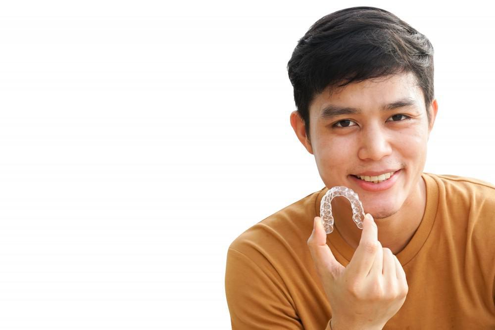 Young man holding up Invisalign retainer while smiling